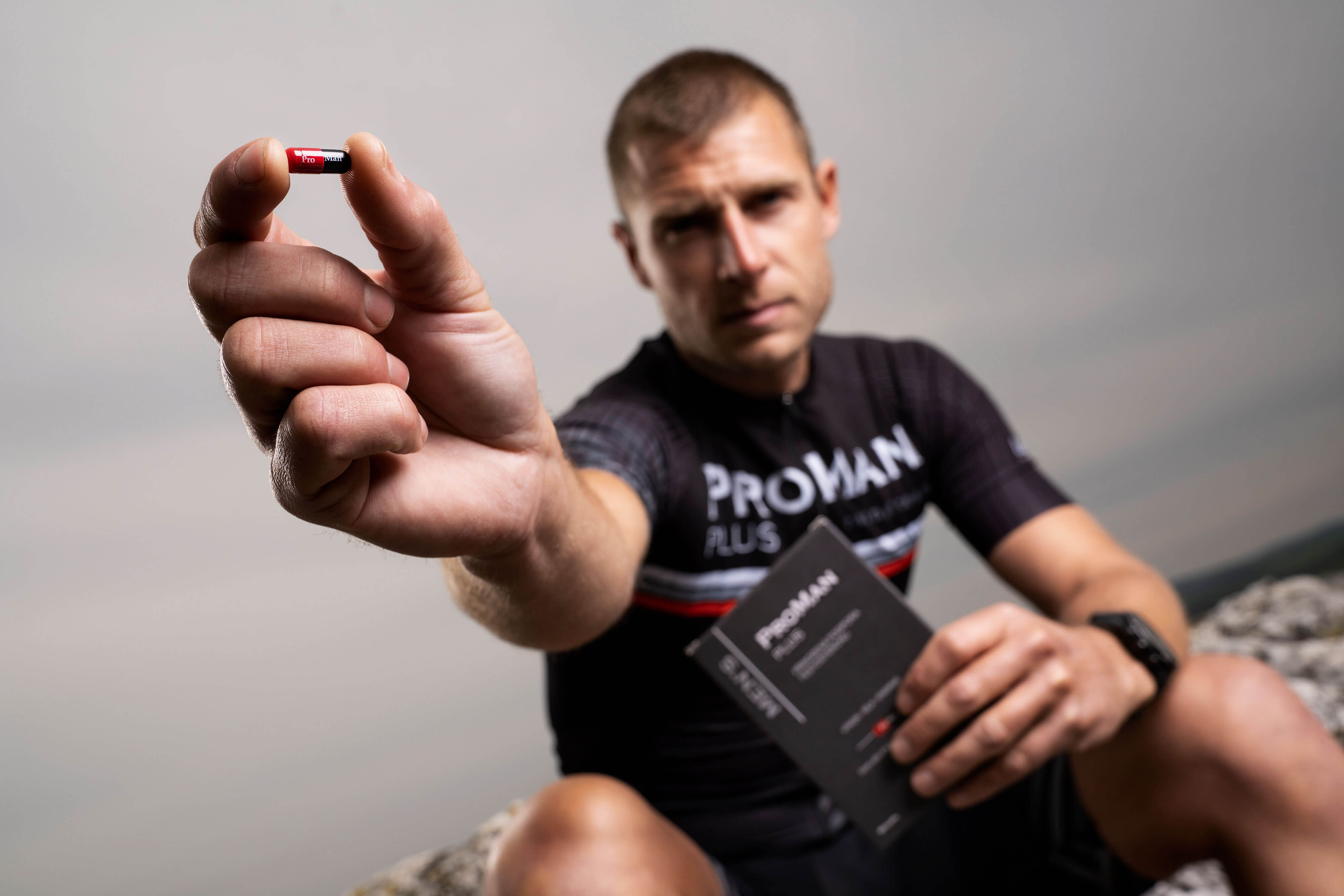 Natural Pharmaceuticals ProMan® Plus Sportlikus riietuses mees, kelle seljal on kiri „ProMan Plus”, hoiab väljasirutatud käes ProMan Plus toidulisandi kapslit, näidates seda esimesel plaanil. Teises käes hoiab ta toote pakendit.