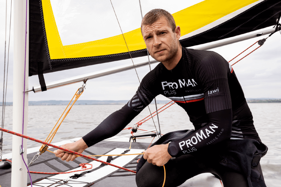 Natural Pharmaceuticals ProMan® Plus Mees spordirõivais kirjaga „ProMan Plus racing team” purjetab jahtil, hoides kinni köiedest ja keskendudes purjede manööverdamisele.